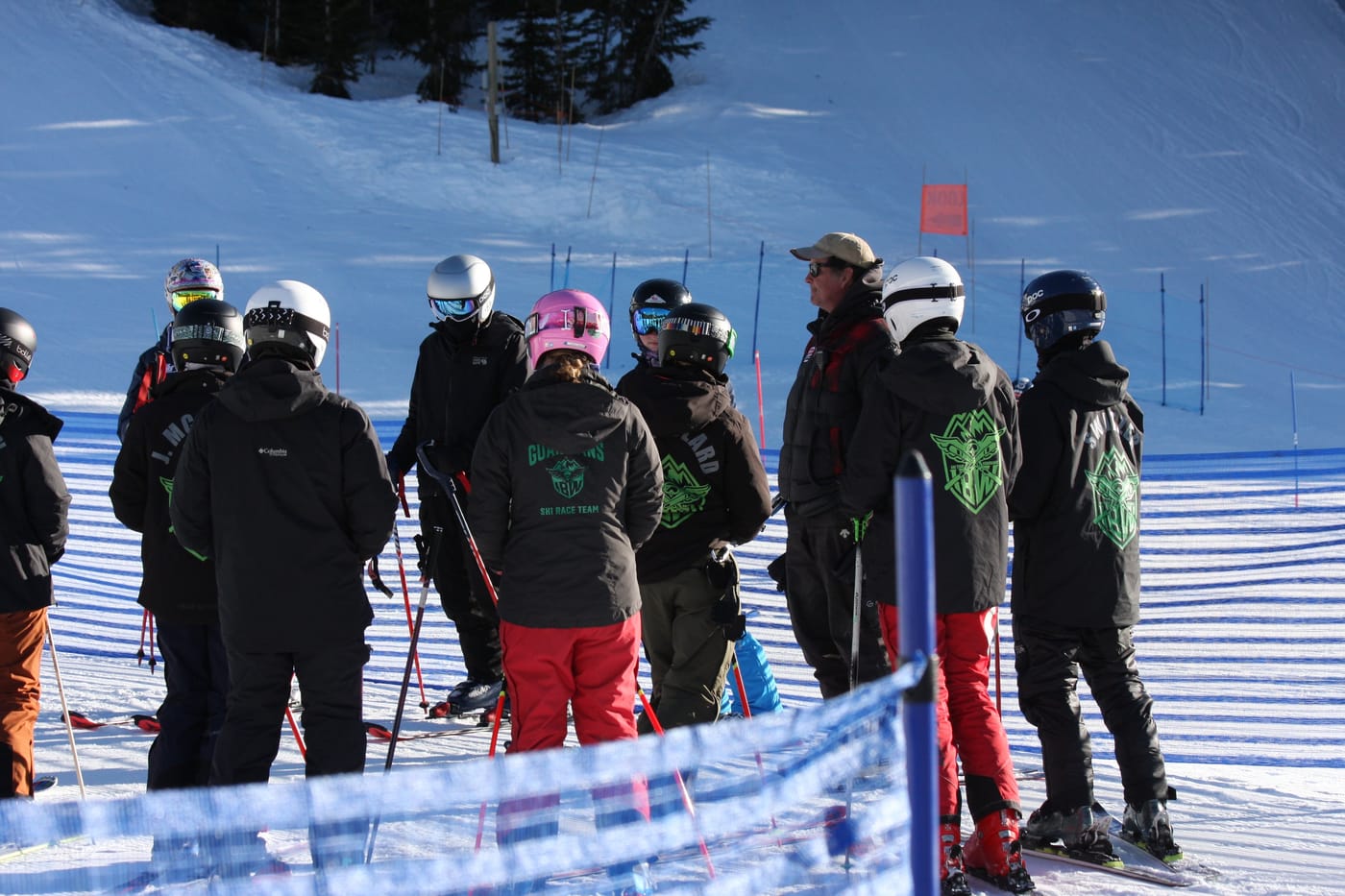 A coach in the middle of the team on the hill, everyone leaning in, the course fence behind them