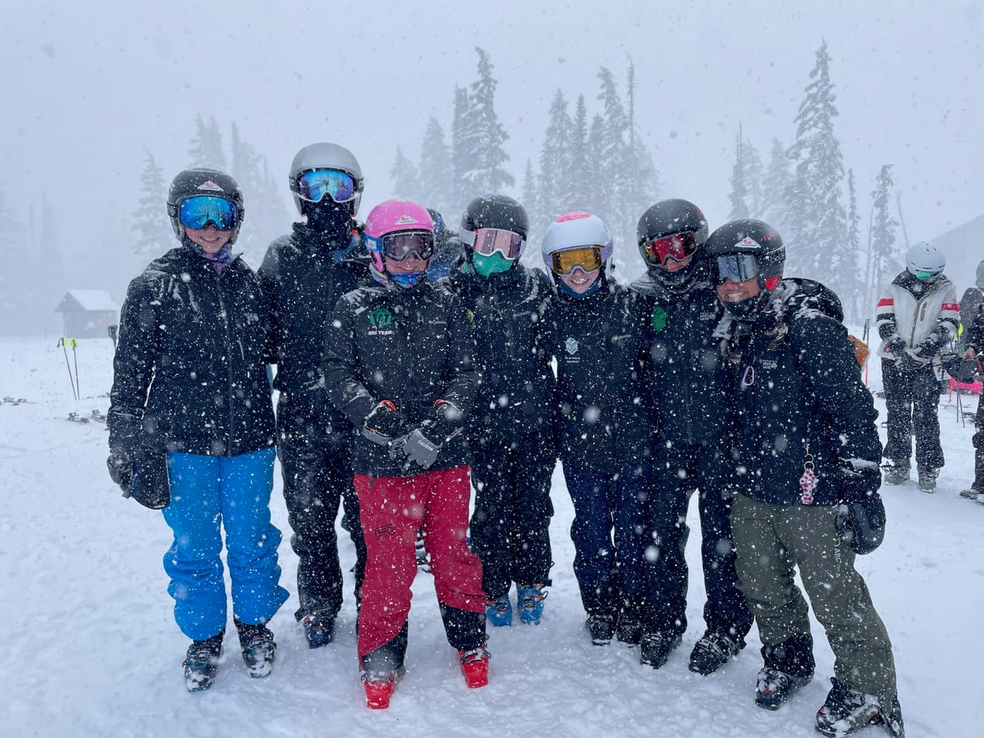 Seven racers packed together and laughing in heavy falling snow, visibility down to the nearest trees